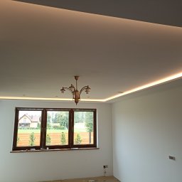 KRISBUD KRZYSZTOF DOROTA STASZEK - Jasne wnętrze z oknem na ogród, widoczne listwy LED pod sufitem i mosiężny żyrandol, świeżo pomalowane ściany w odcieniach bieli, podłoga zabezpieczona folią malarską.