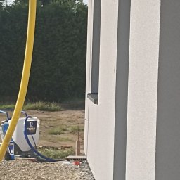 KRISBUD KRZYSZTOF DOROTA STASZEK - Natryskowa aplikacja świeżej, jasnoszarej elewacji na ścianie domu jednorodzinnego, widoczny agregat malarski Graco i żółty wąż doprowadzający farbę, tło stanowią krzewy i trawa.