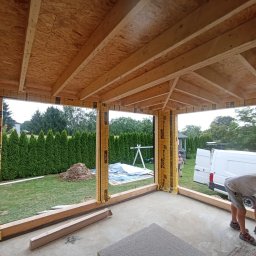 KRISBUD KRZYSZTOF DOROTA STASZEK - Wnętrze w stanie surowym domu w Rudołtowicach, widoczne drewniane elementy konstrukcyjne dachu i ścian obłożonych folią izolacyjną, na zewnątrz widoczny ogród i pracownik przy pracy.