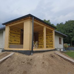 KRISBUD KRZYSZTOF DOROTA STASZEK - Surowy stan rozbudowy domu z widoczną konstrukcją drewnianą i izolacją Eurovent, ziemny nasyp przed budynkiem, pochmurne niebo w tle.