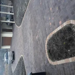 Ułożona kostka brukowa w odcieniach brązu i szarości, tworząca podjazd i ścieżkę z ozdobnymi obrzeżami rabat kwiatowych, widok z góry na zaparkowany samochód i wiatę garażową.