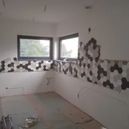 Układanie heksagonalnych płytek w odcieniach szarości na ścianie w kuchni, widoczne niedokończone fragmenty i wystające przewody elektryczne, pod oknem zabezpieczonym niebieską taśmą malarską.