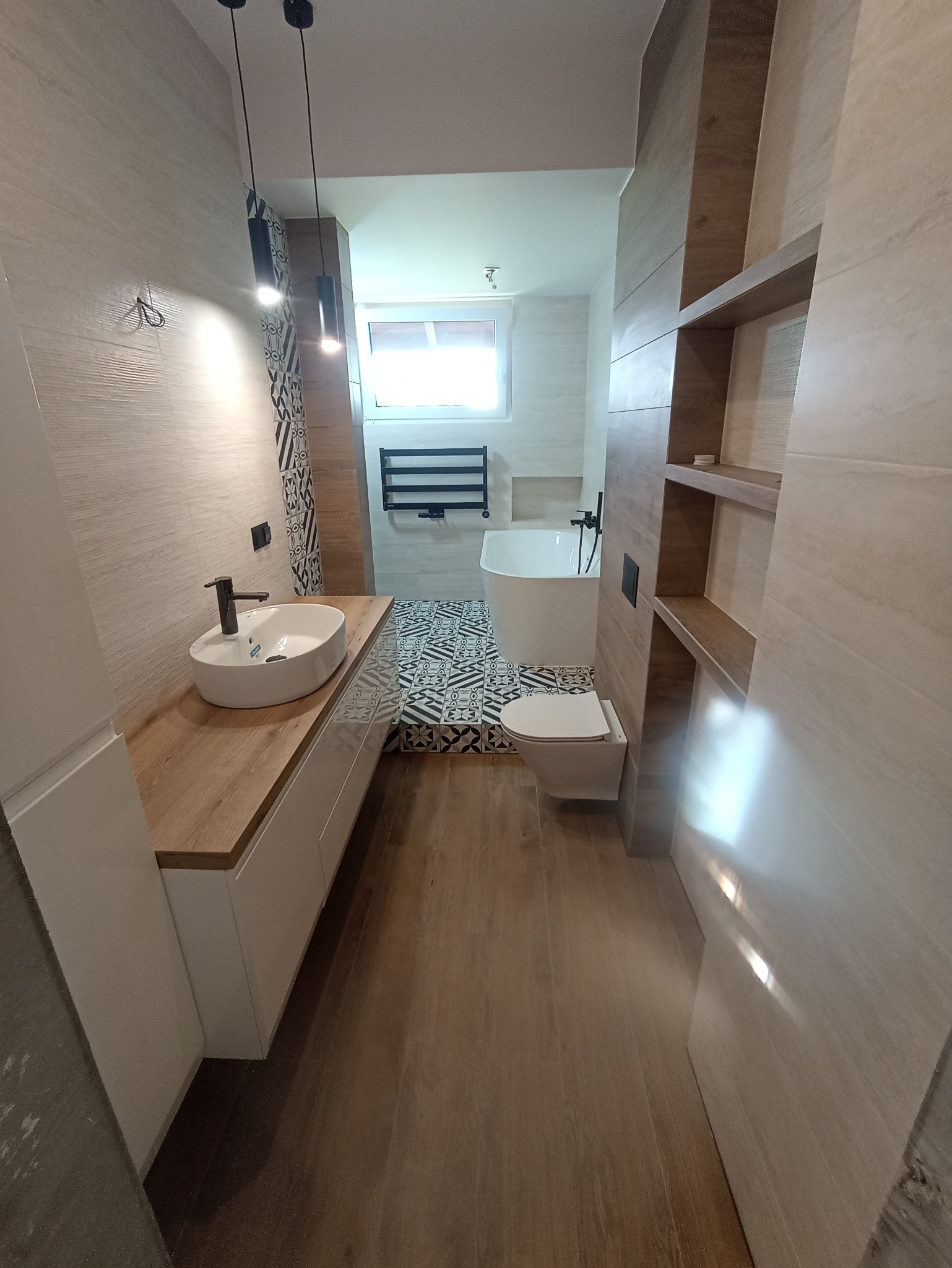 Nowoczesna łazienka z umywalką nablatową na drewnianym blacie, białą wanną wolnostojącą, toaletą i grzejnikiem drabinkowym, wykończona płytkami w odcieniach beżu i brązu z elementami dekoracyjnymi.