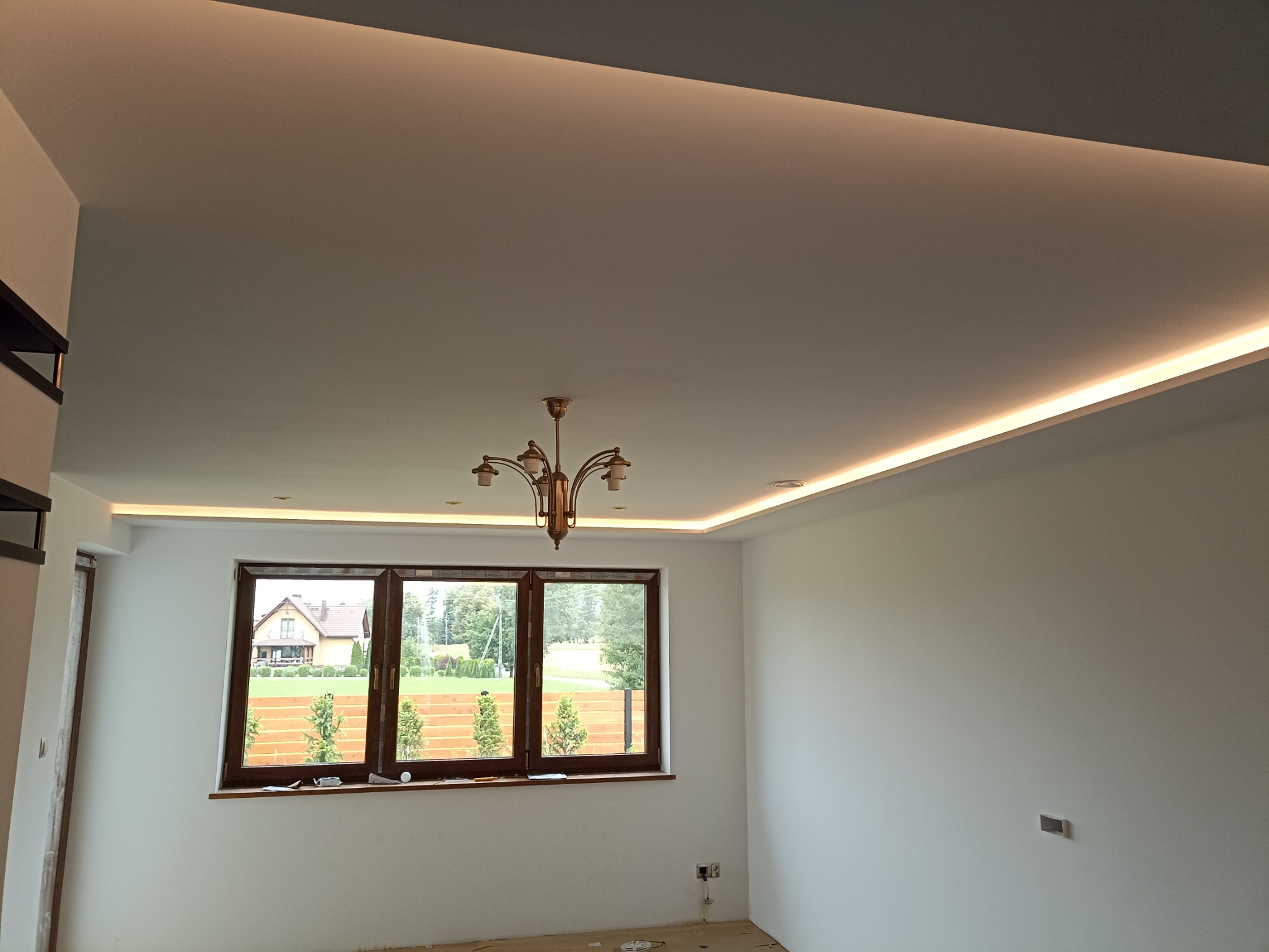 Jasne wnętrze z oknem na ogród, widoczne listwy LED pod sufitem i mosiężny żyrandol, świeżo pomalowane ściany w odcieniach bieli, podłoga zabezpieczona folią malarską.