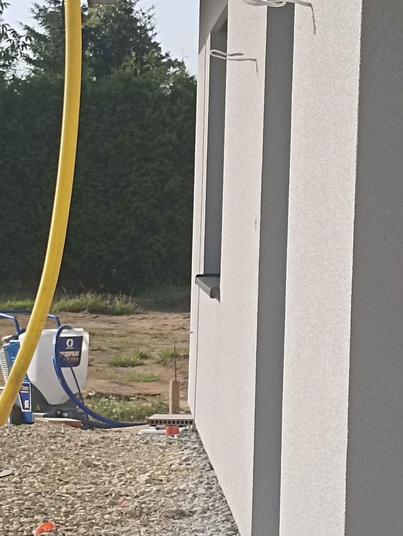 Natryskowa aplikacja świeżej, jasnoszarej elewacji na ścianie domu jednorodzinnego, widoczny agregat malarski Graco i żółty wąż doprowadzający farbę, tło stanowią krzewy i trawa.