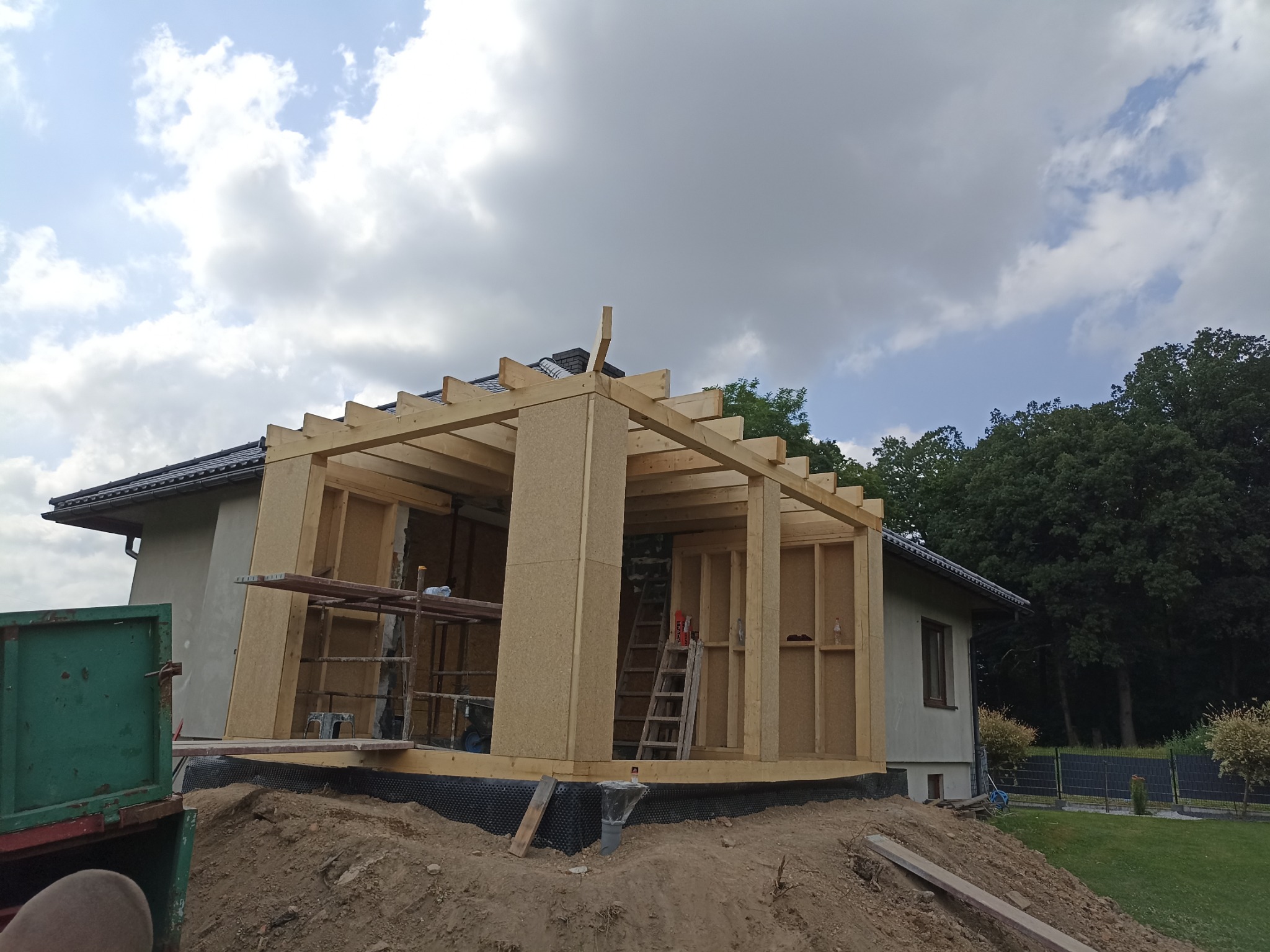 Konstrukcja szkieletowa dobudowy domu z płyty OSB, widok z zewnątrz, etap budowy, drabina oparta o ścianę, ziemia i częściowo widoczny budynek w tle.