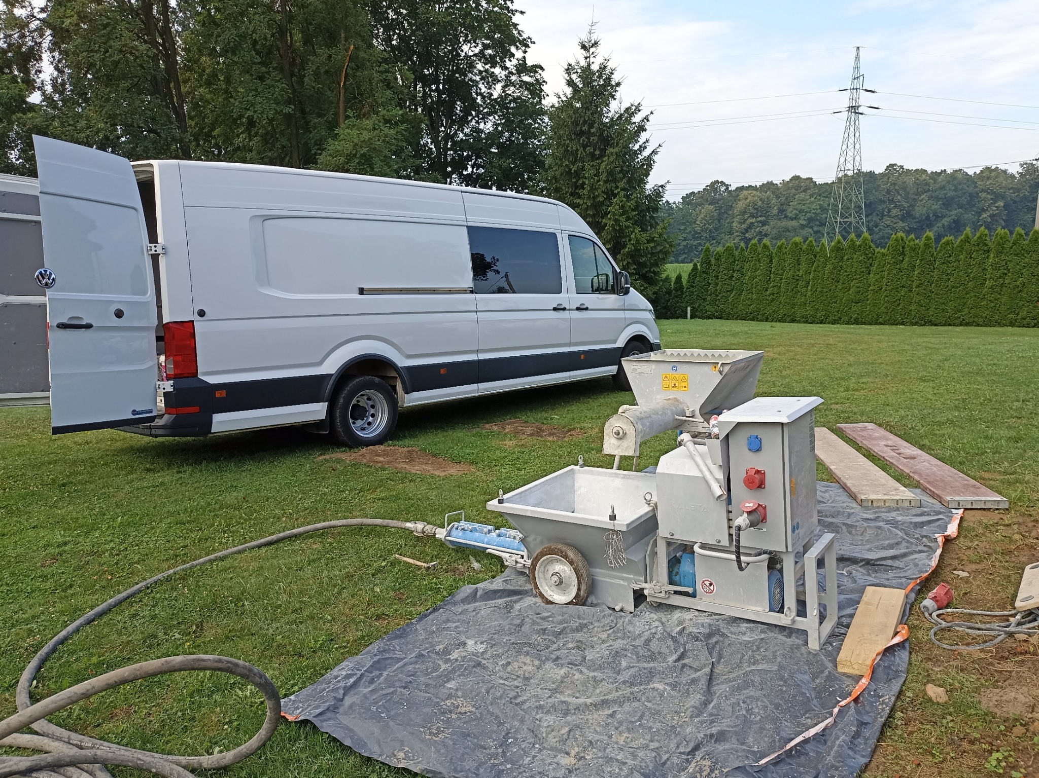 Agregat tynkarski na trawie, z otwartym białym vanem w tle, przygotowany do pracy na zewnątrz.