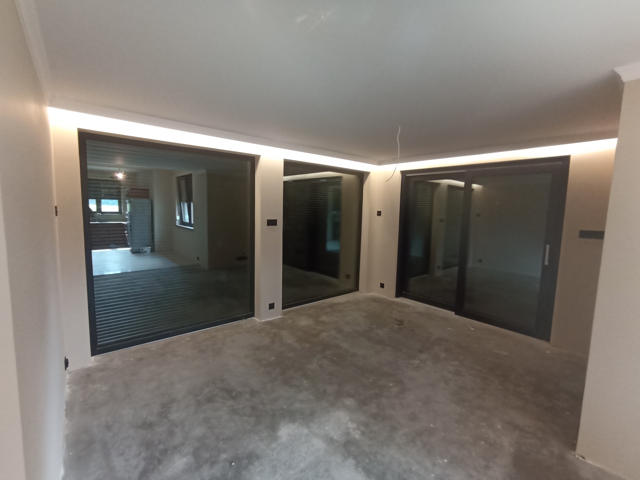 Pusty pokój z trzema dużymi oknami przesuwnymi z czarnymi ramami, surowa betonowa podłoga, oświetlenie LED wzdłuż krawędzi sufitu, widok na sąsiednie pomieszczenie w tle.