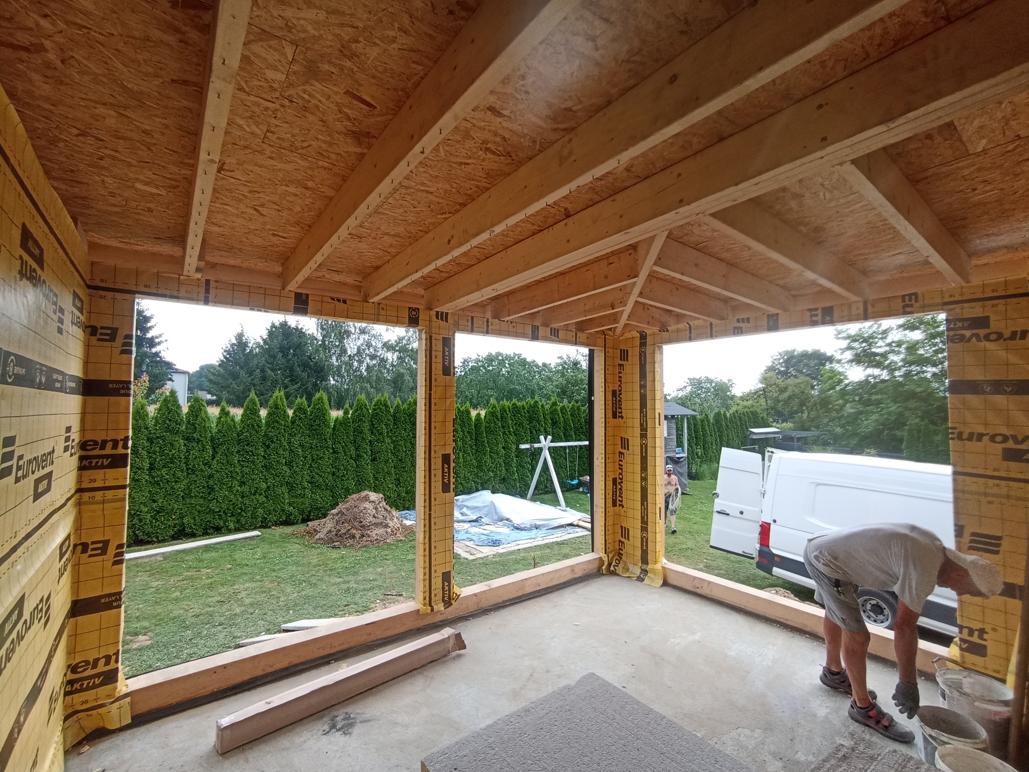 Wnętrze w stanie surowym domu w Rudołtowicach, widoczne drewniane elementy konstrukcyjne dachu i ścian obłożonych folią izolacyjną, na zewnątrz widoczny ogród i pracownik przy pracy.