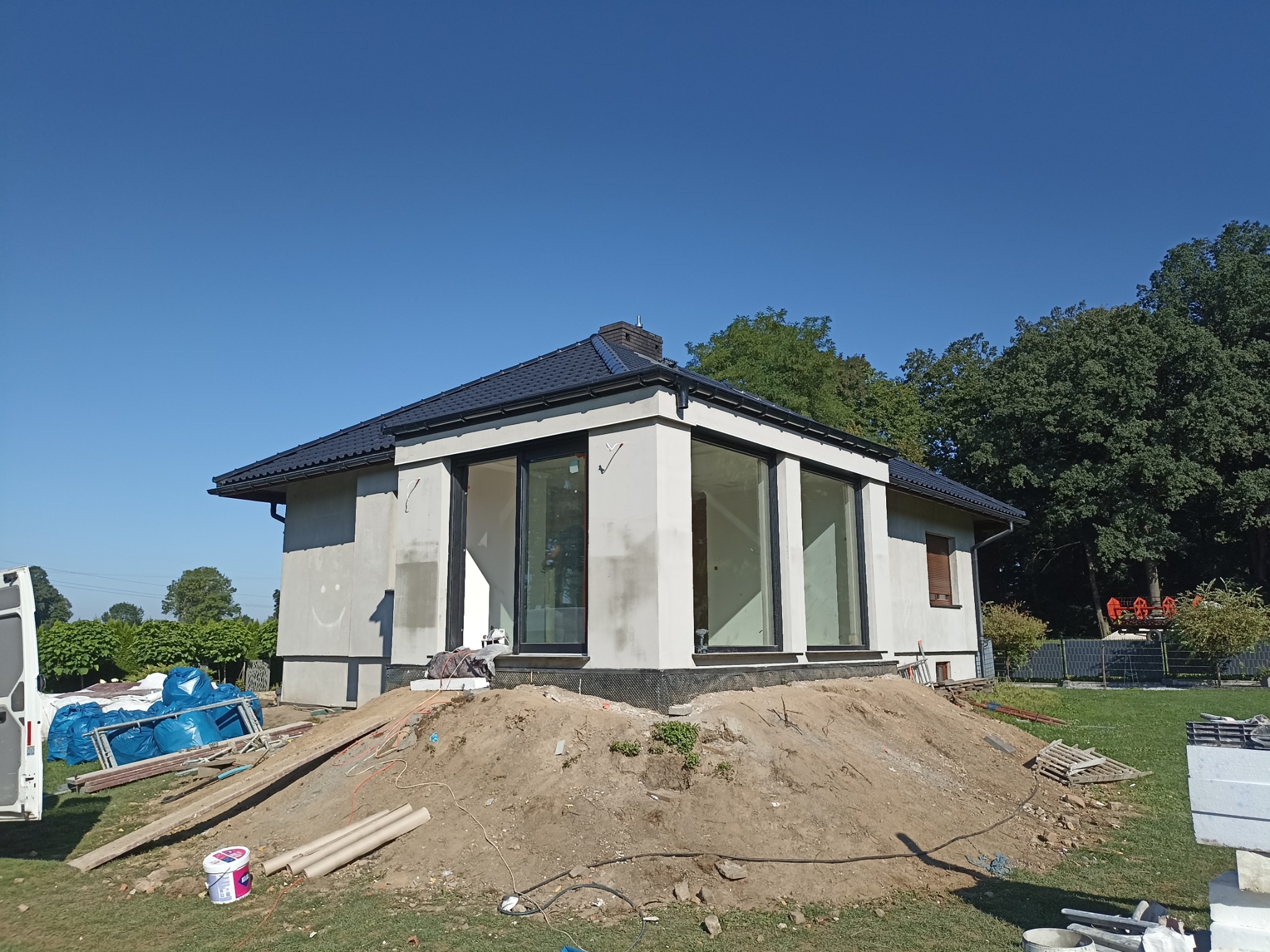 Niedokończony dom jednorodzinny z czarną dachówką i dużymi oknami, otoczony stertą ziemi i materiałami budowlanymi, na ścianie narysowany uśmiech.