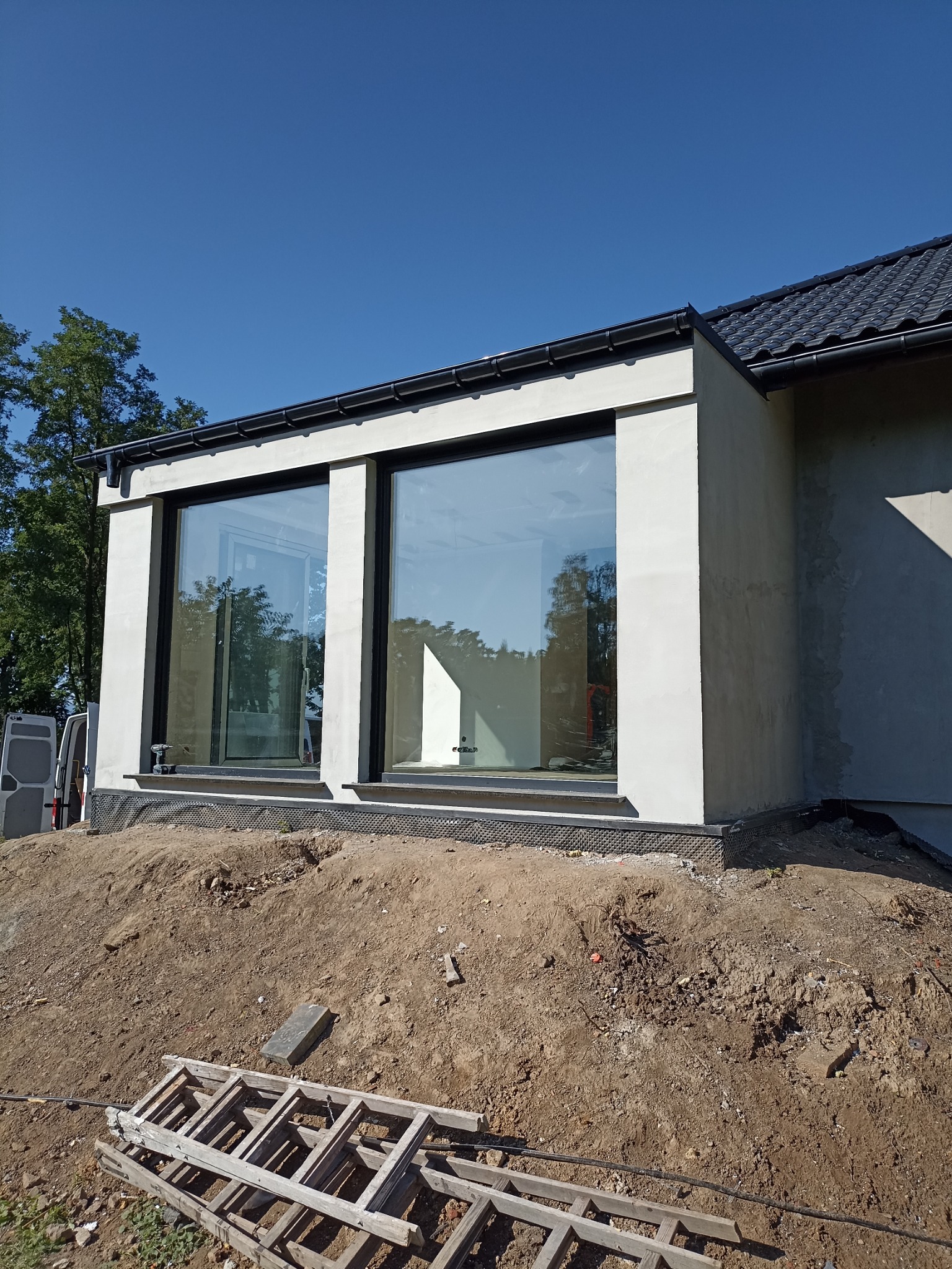 Dobudówka domu z dużymi oknami w czarnych ramach, widoczny fragment dachu z dachówką oraz ziemia z resztkami materiałów budowlanych i drabina.