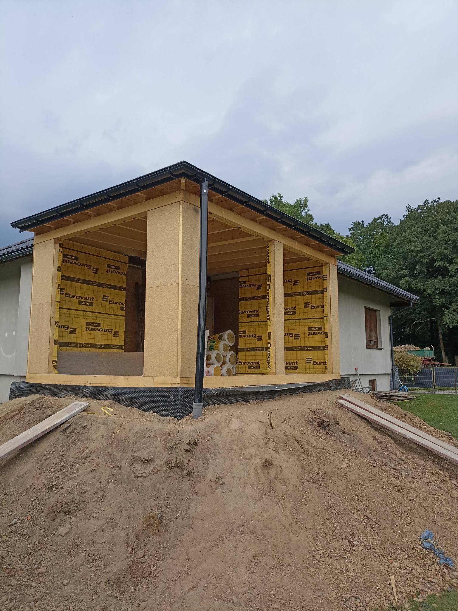 Surowy stan rozbudowy domu z widoczną konstrukcją drewnianą i izolacją Eurovent, ziemny nasyp przed budynkiem, pochmurne niebo w tle.