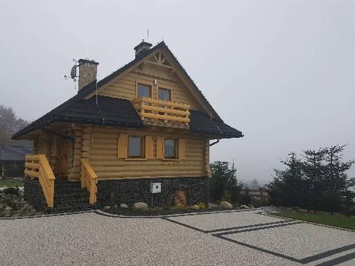 Dwukondygnacyjny dom z bali z balkonem, czarnym dachem i kamienną podmurówką, otoczony szarą kostką brukową i mglistym krajobrazem.