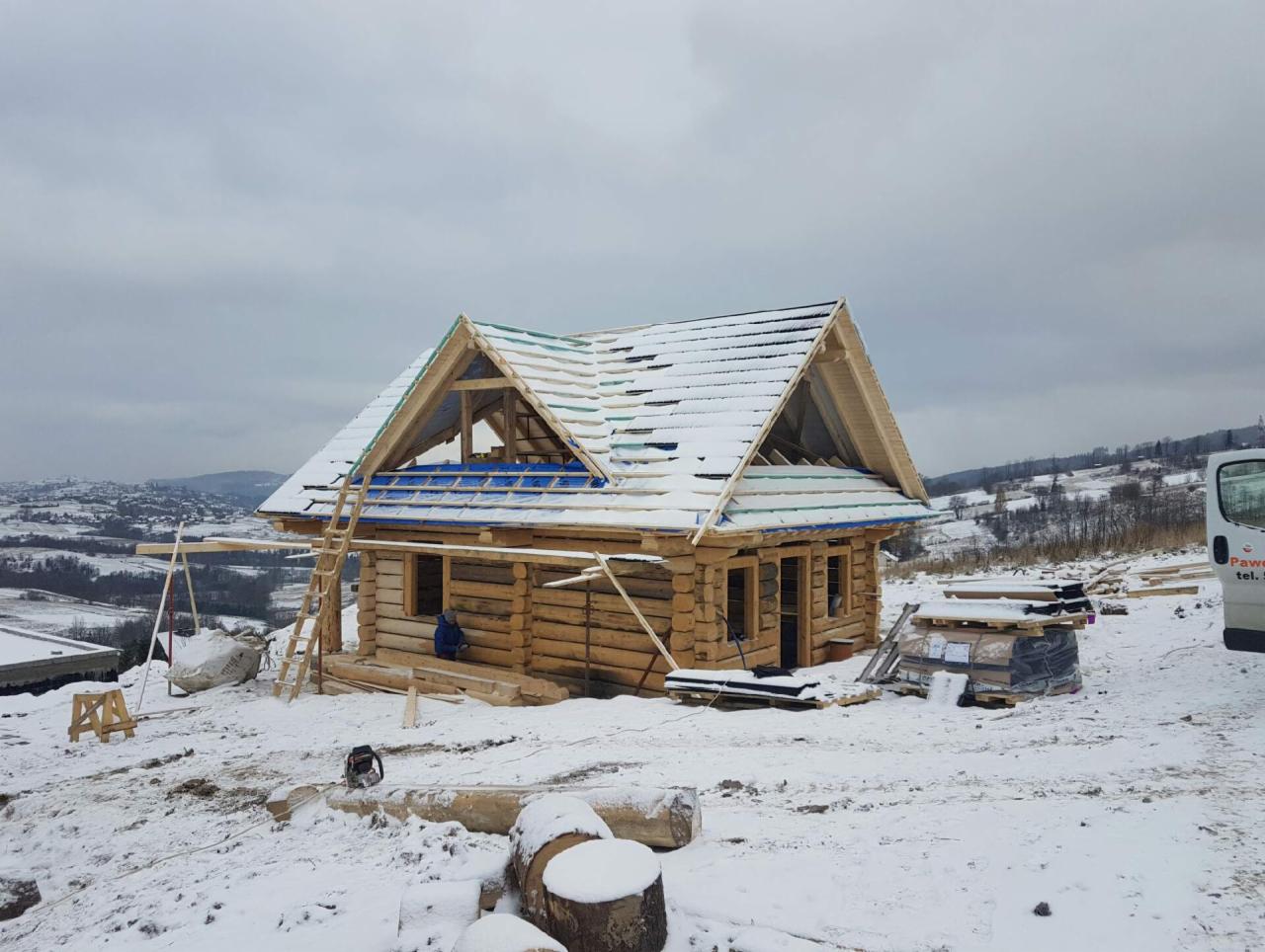 Budowa domu szkieletowego z bali w zimowej scenerii, widoczne rusztowania, częściowo ośnieżony dach i okoliczne wzgórza.