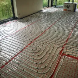 Białe rury ogrzewania podłogowego ułożone na folii z czerwoną siatką, widoczne okno z zielenią na zewnątrz oraz kartony z napisem 'Terma Organika'.