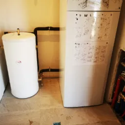 Biały zbiornik akumulacyjny ciepłej wody użytkowej obok pompy ciepła Vaillant z naklejoną instrukcją, połączone czarnymi rurami w pomieszczeniu technicznym z szarą podłogą z płytek.