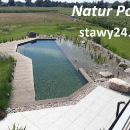 Staw kąpielowy z drewnianym pomostem i strefą roślinną, otoczony trawnikiem i polami uprawnymi, widok z góry z elementami balustrady w prawym dolnym rogu.