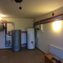 AVM ENGINEERING Sp. z o.o. - Dwa zbiorniki buforowe, szary i biały, w kotłowni z instalacją hydrauliczną z rur miedzianych i niebieskich, widoczne także grzejniki i elementy sterujące.