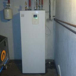 AVM ENGINEERING Sp. z o.o. - Biała jednostka wewnętrzna pompy ciepła w piwnicy, widoczne rury instalacji, manometr, zbiornik wyrównawczy i fragment wózka narzędziowego.
