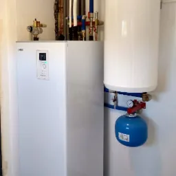 Biała jednostka wewnętrzna NIBE z podłączonym zasobnikiem ciepłej wody i niebieskim naczyniem przeponowym, widoczne rury instalacji grzewczej w tle.