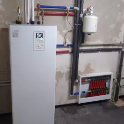 Biała jednostka centralna z wyświetlaczem, podłączona do rur w kolorze niebieskim i czerwonym, z widocznym manometrem i zbiornikiem wyrównawczym, na tle surowej ściany i ciemnej podłogi.