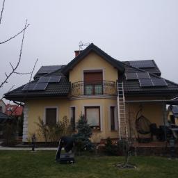 AVM ENGINEERING Sp. z o.o. - Żółty dom jednorodzinny z ciemną dachówką, na której zamontowane są panele słoneczne; na trawniku stoi mężczyzna w czarnym ubraniu pochylony nad czarną skrzynką narzędziową, obok domu przystawiona...