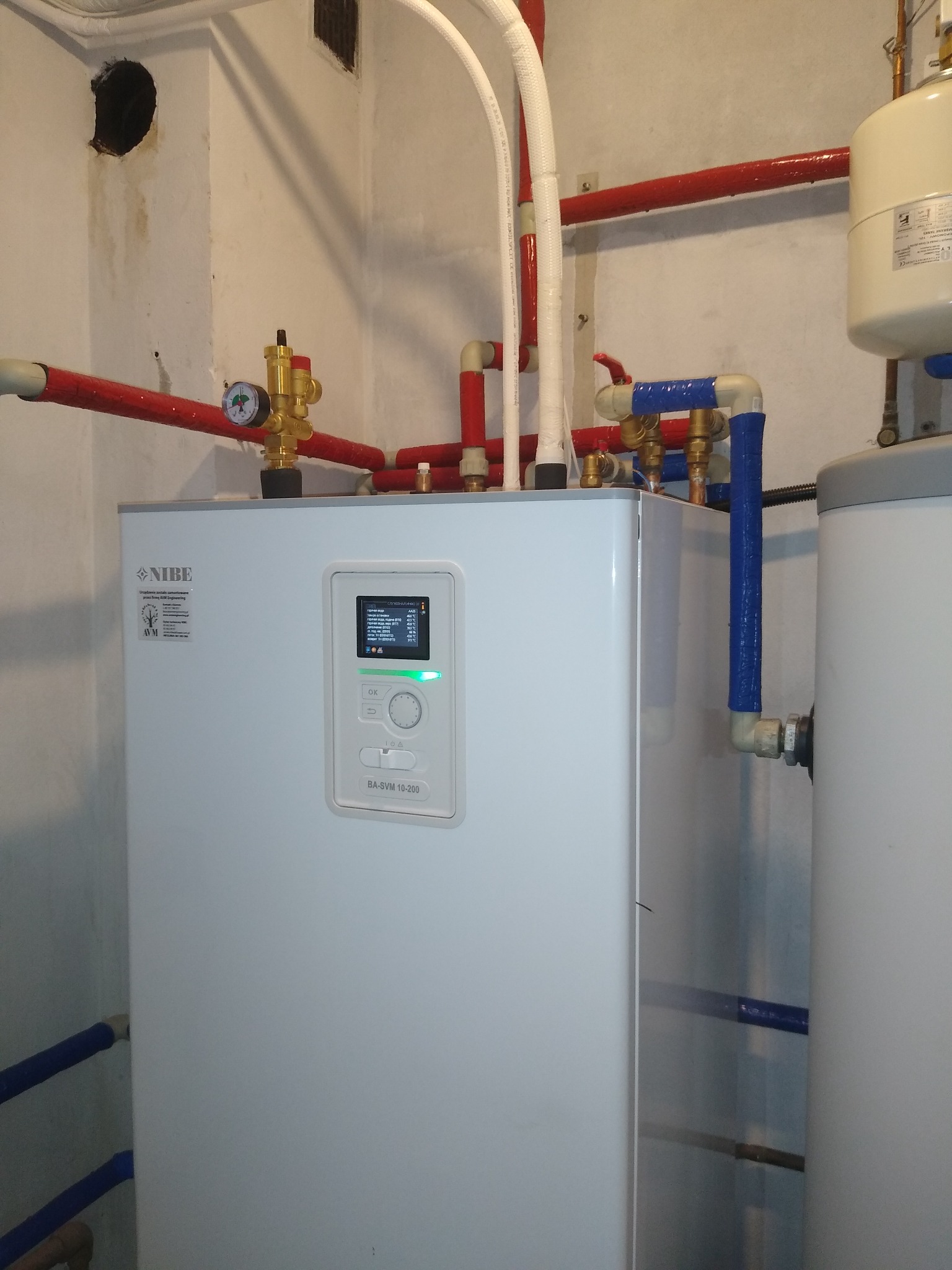 Biała jednostka centralna NIBE BA-SVM 10-200 z ekranem wyświetlającym parametry pracy, otoczona instalacją hydrauliczną z czerwonymi i niebieskimi rurami w pomieszczeniu technicznym.