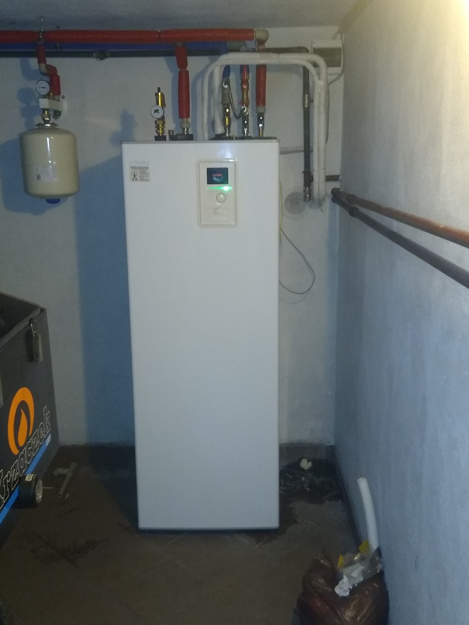 Biała jednostka wewnętrzna pompy ciepła w piwnicy, widoczne rury instalacji, manometr, zbiornik wyrównawczy i fragment wózka narzędziowego.