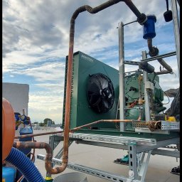 GREEN SERWIS klimatyzacja-chłodnictwo-wentylacja Rafał Zieliński - Montaż przemysłowego agregatu chłodniczego Arkton na dachu, widoczne rury miedziane, wentylator, narzędzia w wiadrze Castorama i błękitne, obłoczne niebo w tle.