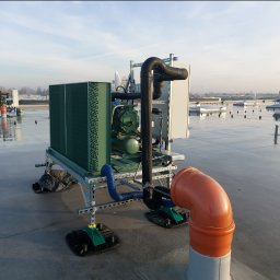 GREEN SERWIS klimatyzacja-chłodnictwo-wentylacja Rafał Zieliński - Agregat chłodniczy na dachu budynku, widoczne rury, przewody i elementy konstrukcyjne, tło stanowi niebo i odległe budynki.