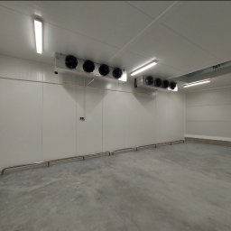 GREEN SERWIS klimatyzacja-chłodnictwo-wentylacja Rafał Zieliński - Wnętrze chłodni przemysłowej z zainstalowanymi agregatami chłodniczymi pod sufitem i zabezpieczeniami ścian. Widoczne białe panele ścienne i betonowa podłoga.