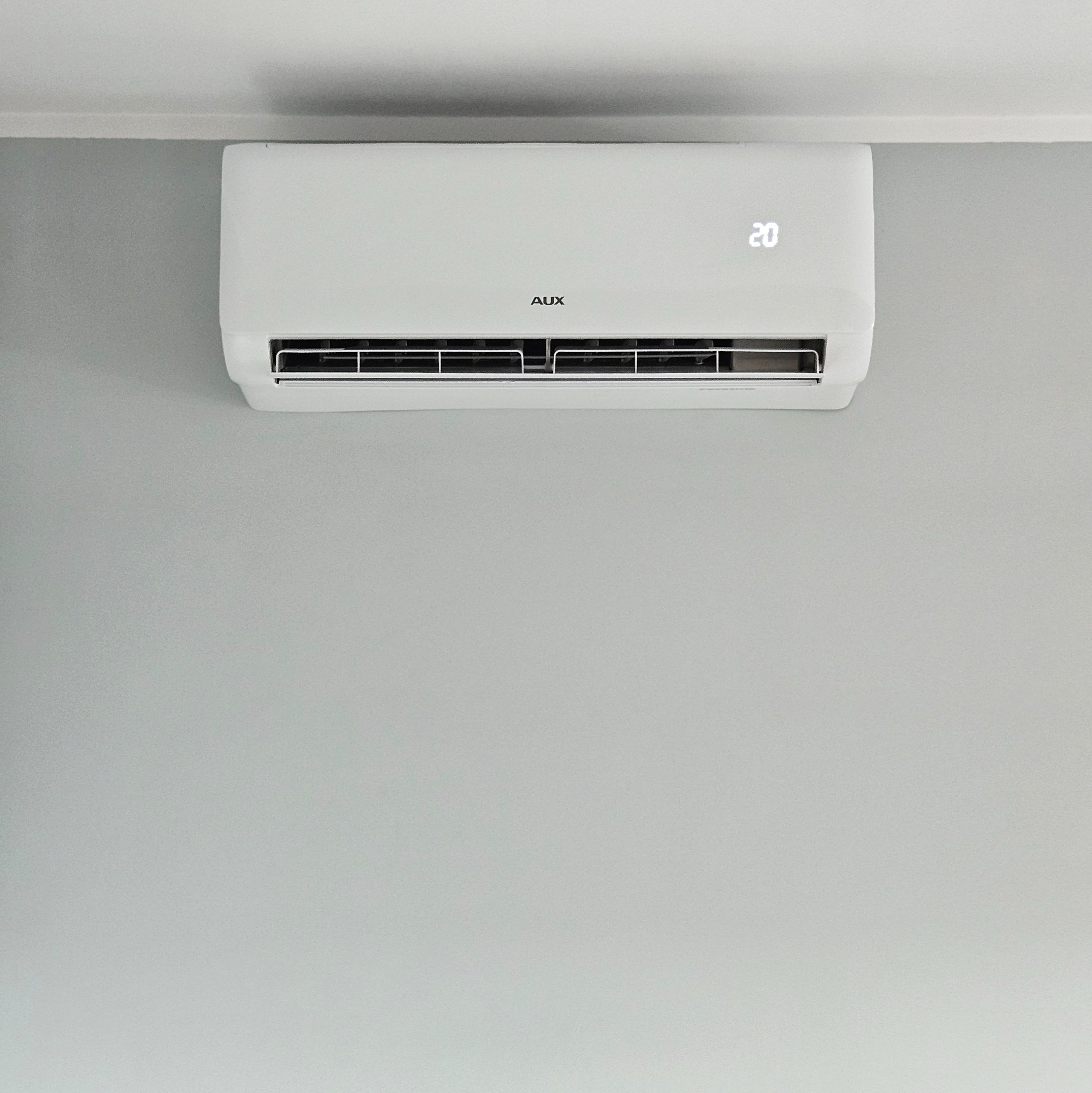 Biały klimatyzator marki AUX zamontowany na szarej ścianie, wyświetlający temperaturę 20 stopni Celsjusza.