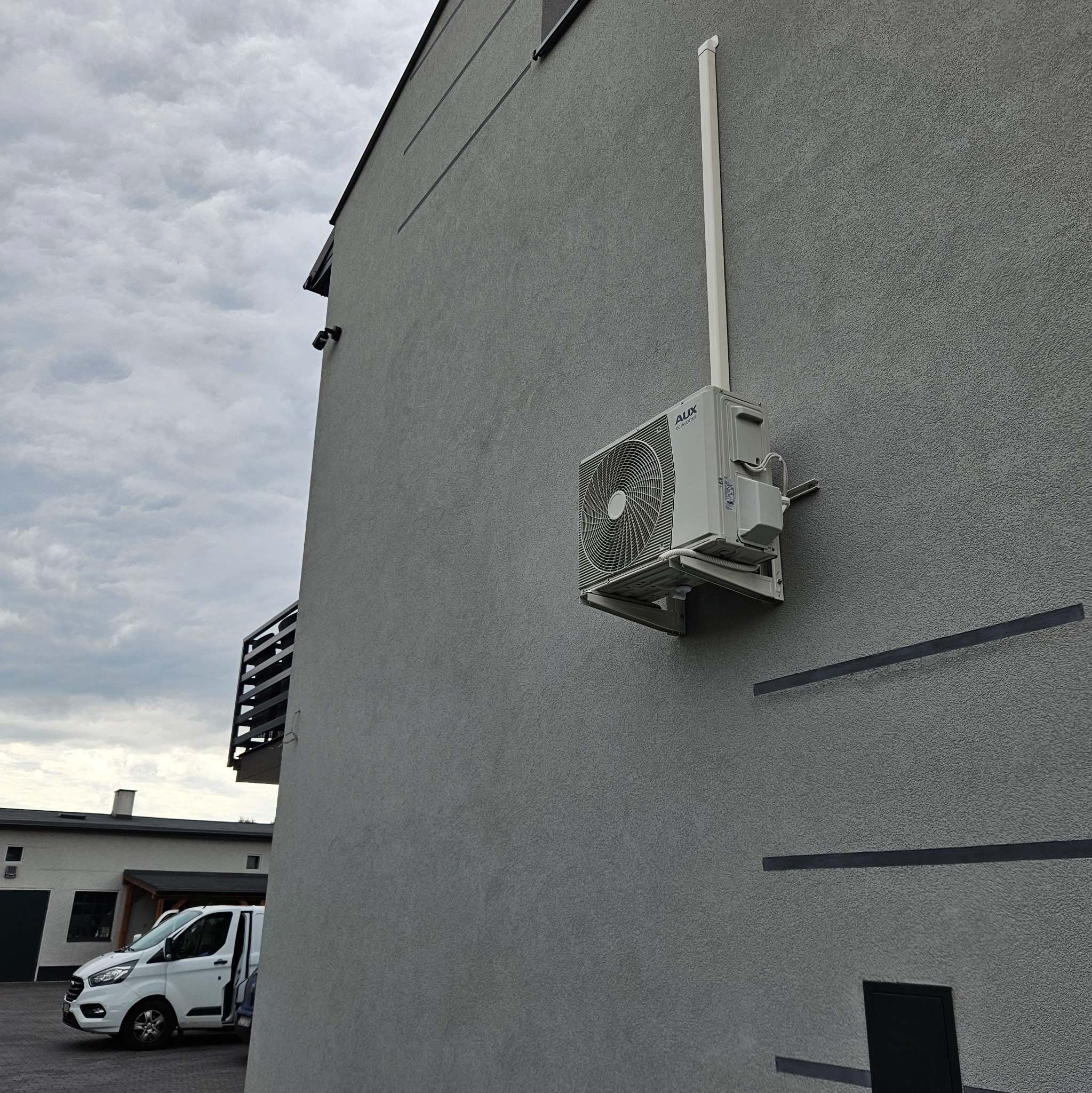 Zewnętrzna jednostka klimatyzacji AUX zamontowana na szarej elewacji budynku, widoczny biały przewód i fragment zaparkowanego białego samochodu dostawczego w tle.