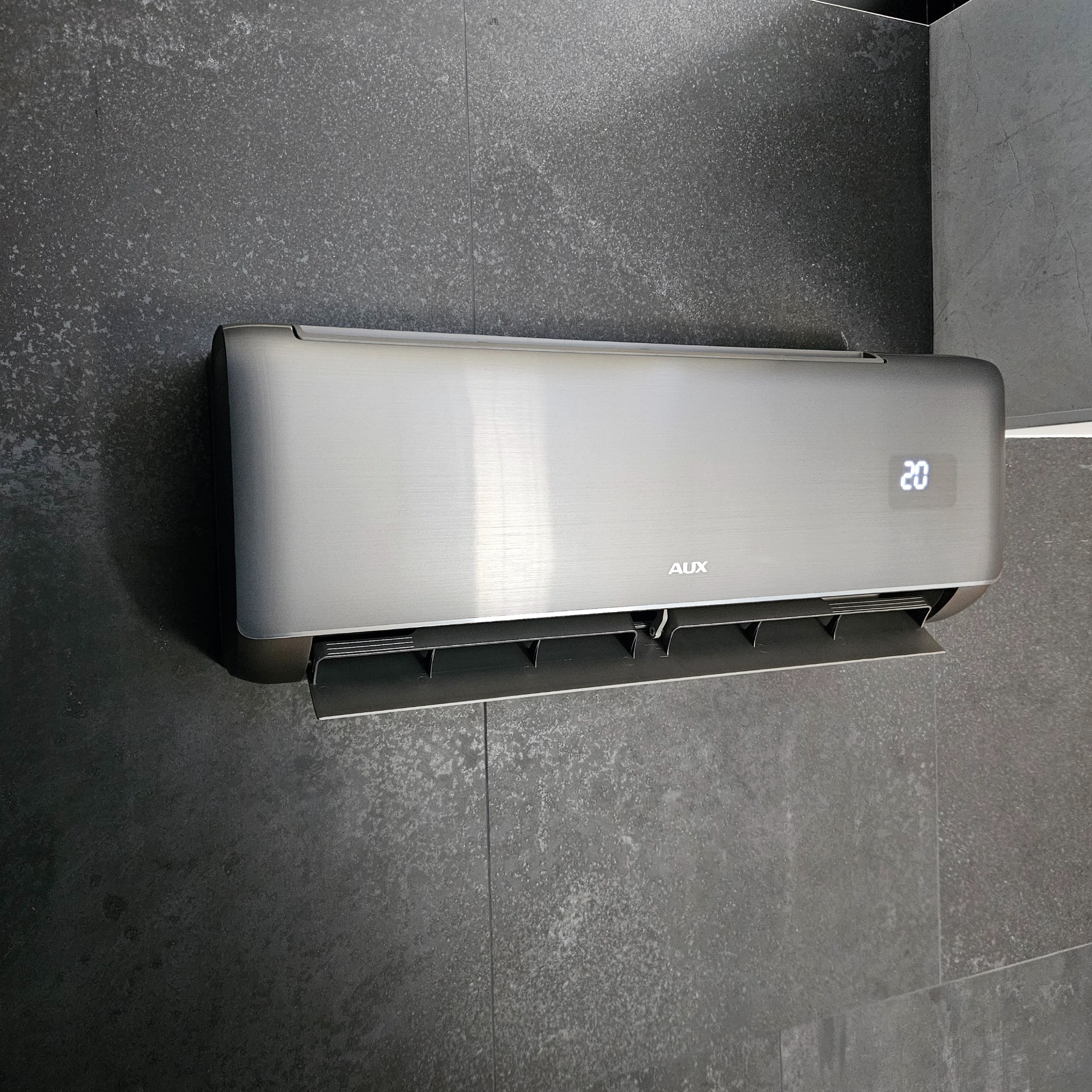 Szary klimatyzator AUX z wyświetlaczem ustawionym na 20 stopni Celsjusza, zamontowany na ścianie wyłożonej ciemnoszarymi płytkami imitującymi kamień.