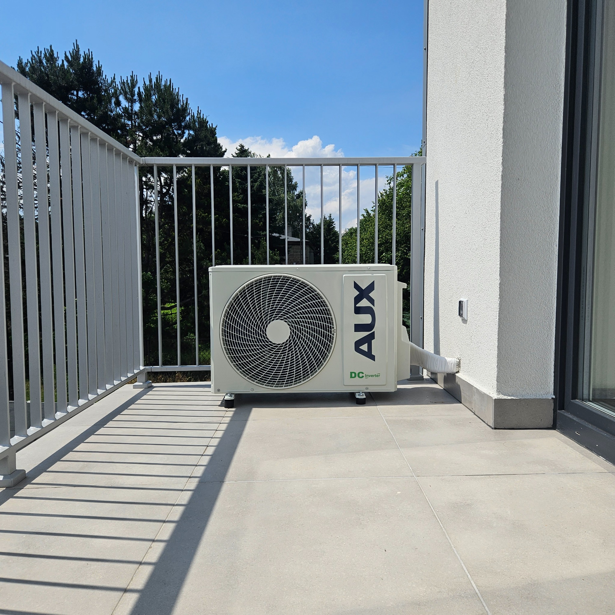Jednostka zewnętrzna klimatyzatora AUX zamontowana na balkonie z widokiem na drzewa i błękitne niebo.