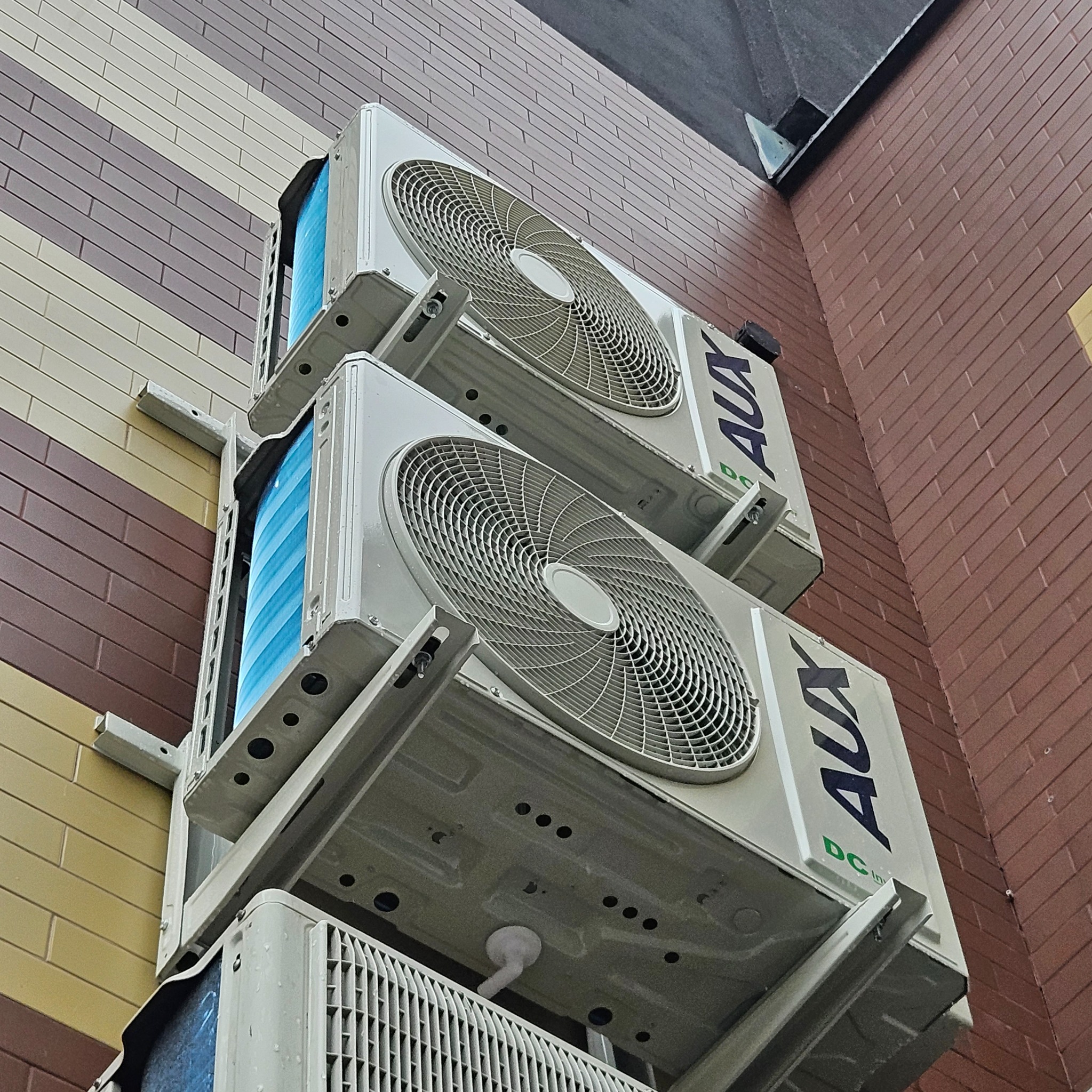 Dwa klimatyzatory AUX zamontowane jeden nad drugim na metalowej konstrukcji na ścianie budynku z cegły i paneli.