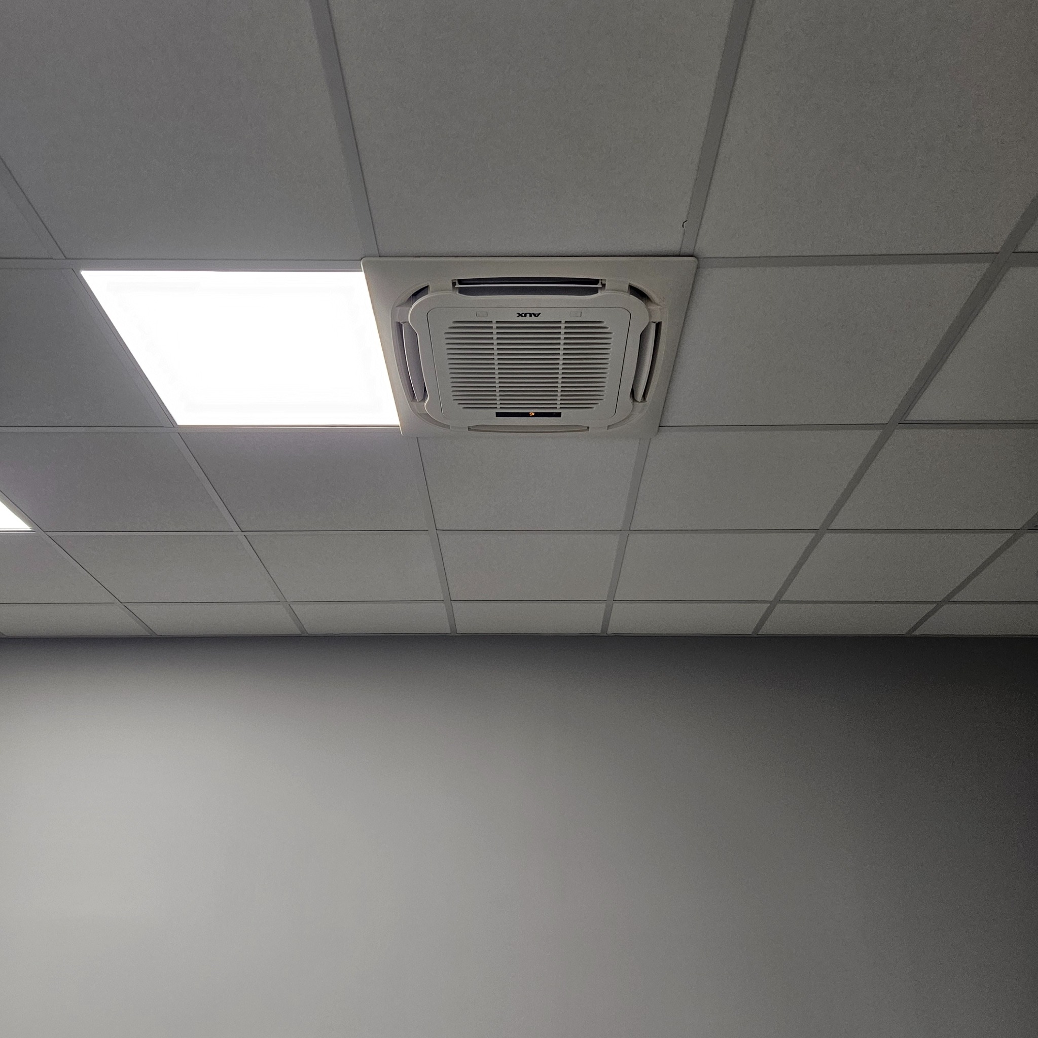 Sufitowa jednostka klimatyzacyjna AUX w kasetonie sufitu podwieszanego, widok z dołu na szarą ścianę.