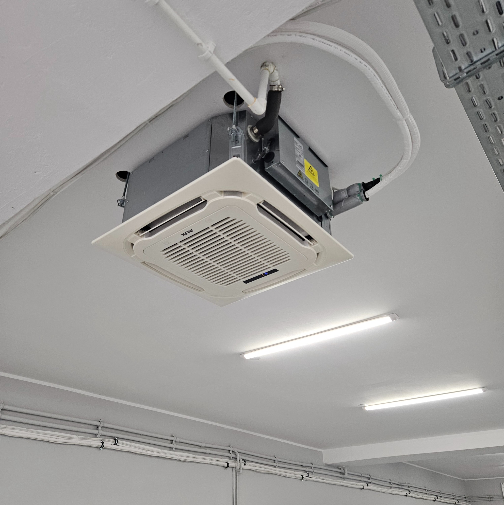 Sufitowa jednostka klimatyzacyjna AUX typu kasetonowego, widoczne rury i instalacja elektryczna, oświetlenie LED.
