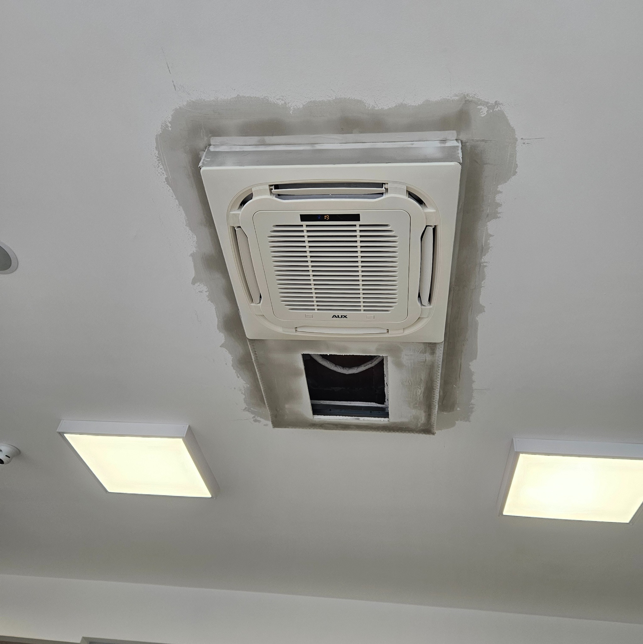 Kasetonowa jednostka klimatyzacyjna AUX zamontowana w suficie, z widocznymi śladami świeżego gładzi szpachlowej wokół obudowy i otwartym otworem instalacyjnym pod spodem.
