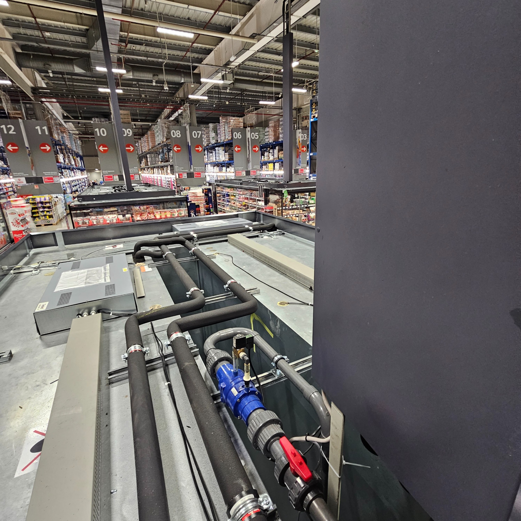 Instalacja rur chłodniczych z zaworami w supermarkecie, widok z góry na system chłodzenia, rury w izolacji, elementy instalacji na tle regałów sklepowych.