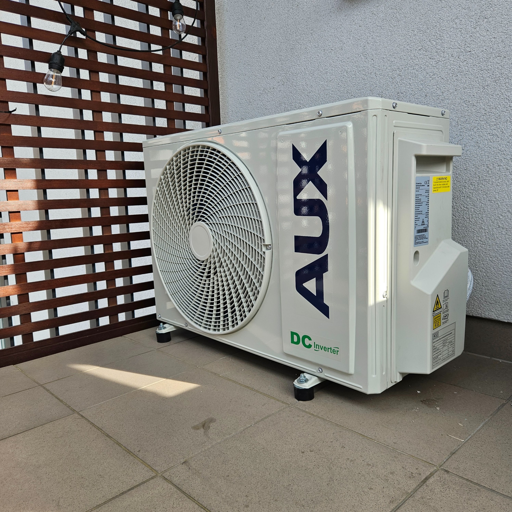 Zewnętrzna jednostka klimatyzacji marki AUX, model DC Inverter, ustawiona na balkonie z drewnianą kratą i dekoracyjnymi żarówkami w tle.