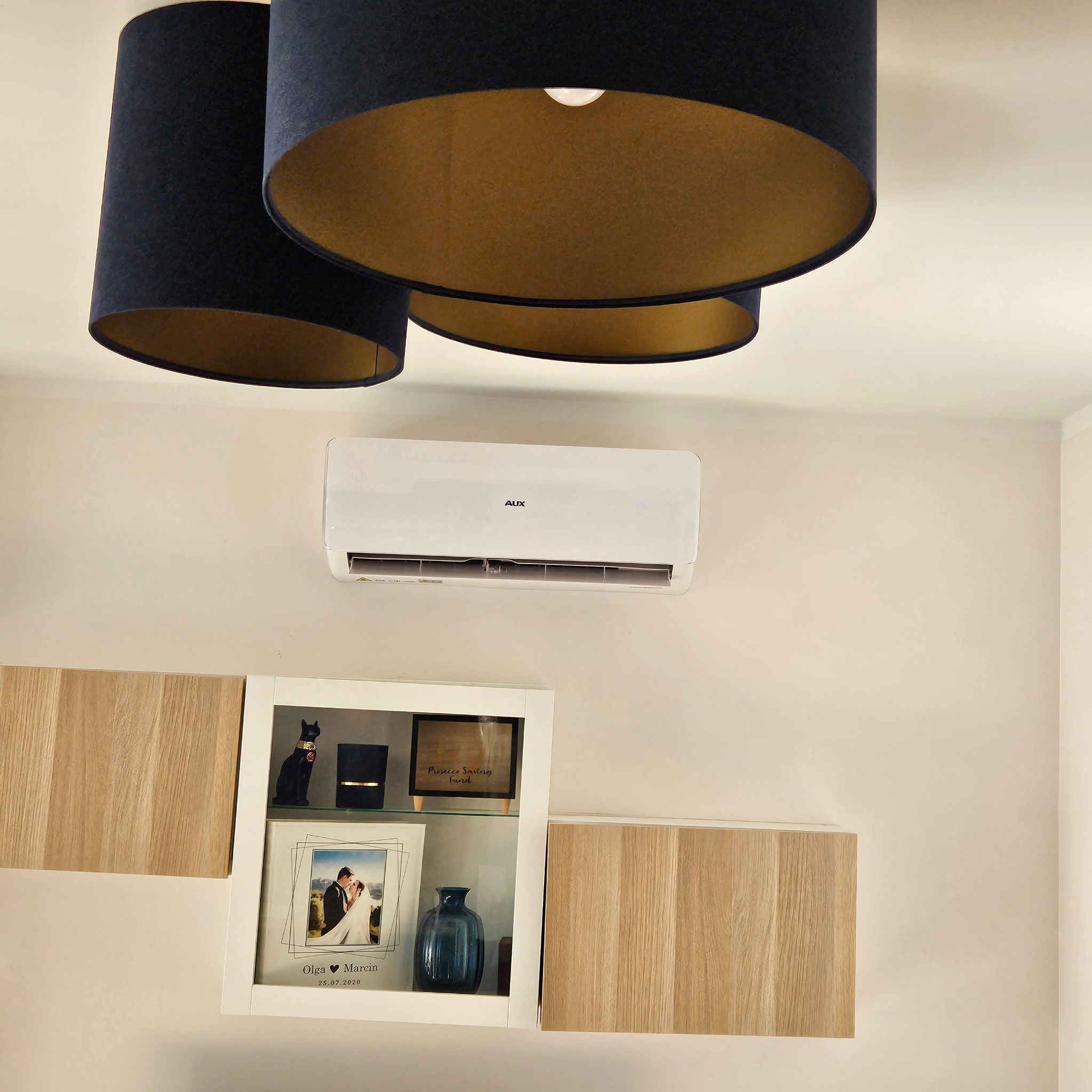 Nowoczesny klimatyzator AUX zamontowany na ścianie w salonie, widoczne lampy z ciemnymi abażurami i jasnymi wnętrzami oraz półki z dekoracjami.