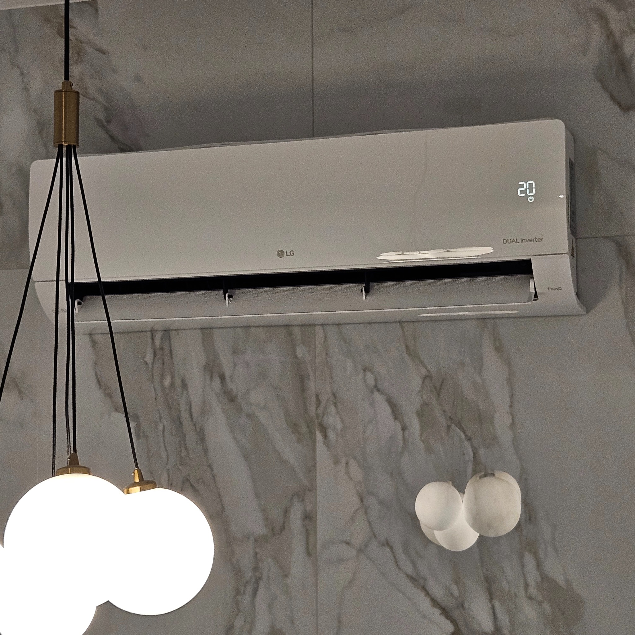 Nowoczesny, biały klimatyzator LG Dual Inverter zamontowany na ścianie wykończonej marmurkowym wzorem, widoczny fragment designerskiej lampy z trzema kulistymi kloszami.