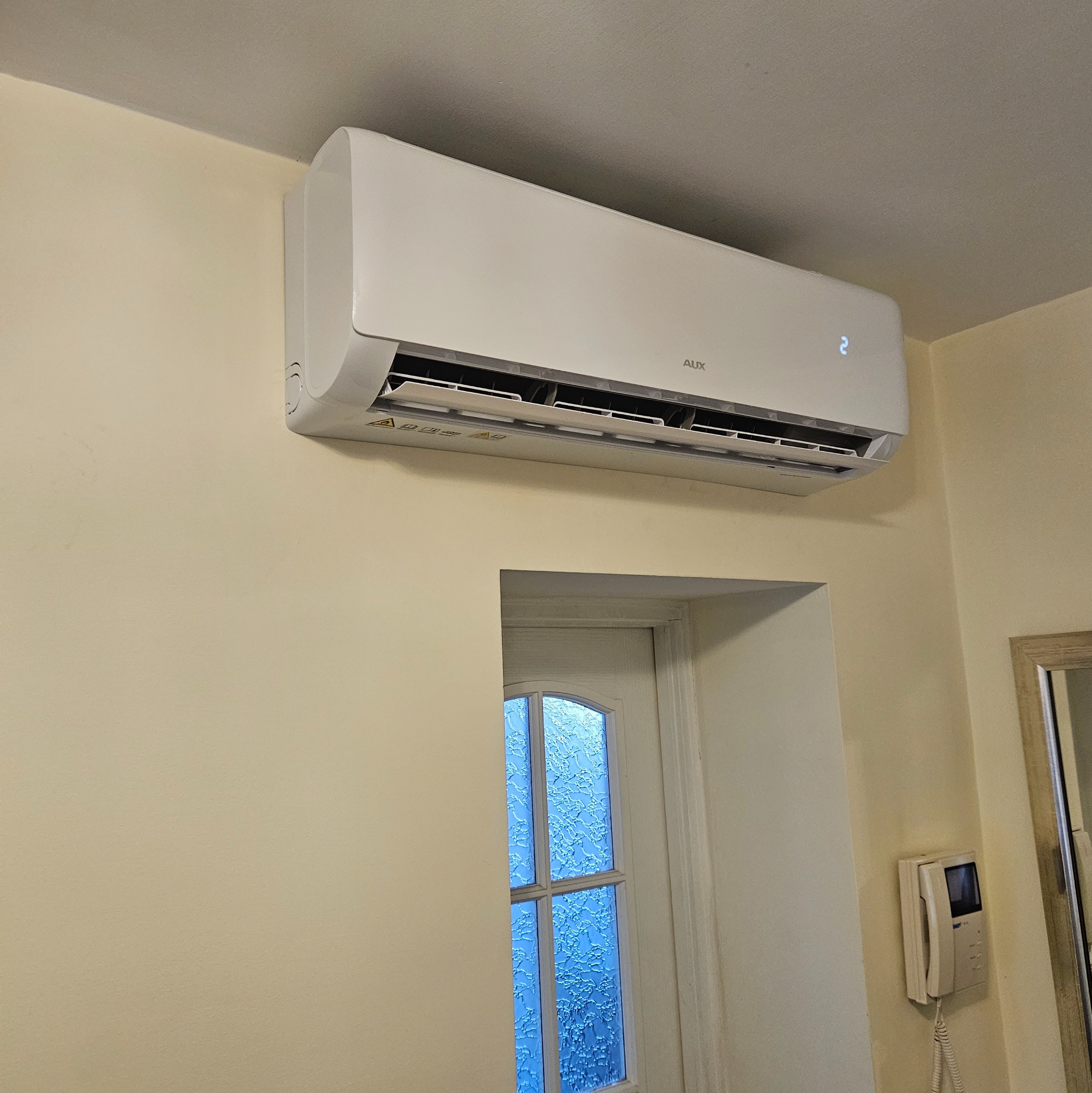 Biała jednostka klimatyzacji AUX zamontowana na ścianie nad oknem z ozdobnym szkłem w jasnym pomieszczeniu.