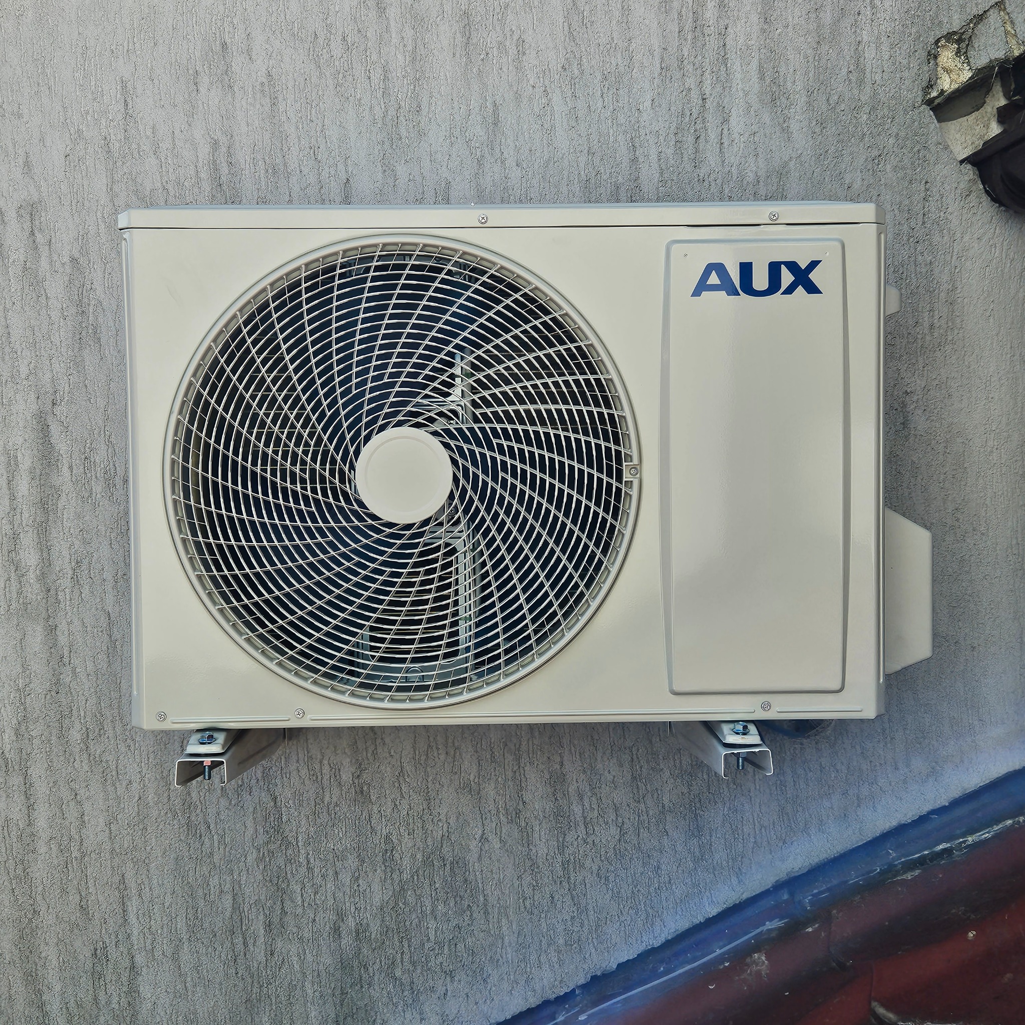 Zewnętrzna jednostka klimatyzatora marki AUX zamontowana na szarej, chropowatej ścianie budynku.
