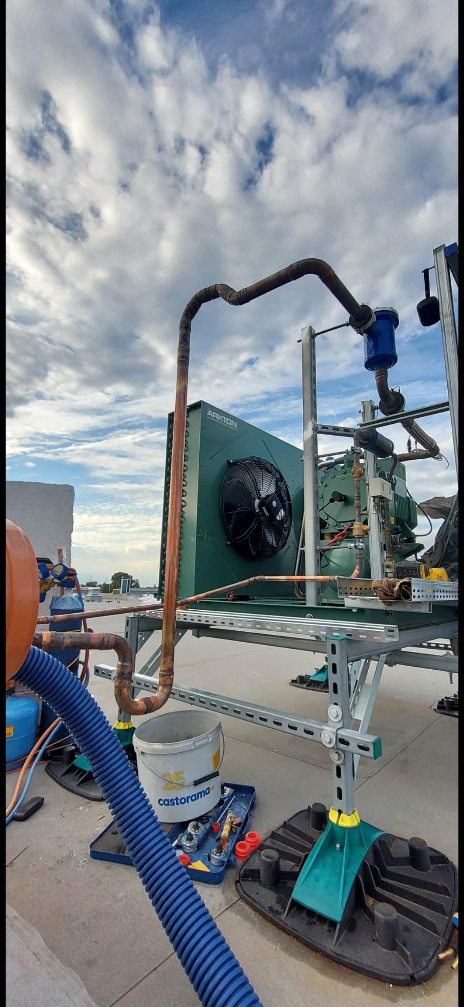 Montaż przemysłowego agregatu chłodniczego Arkton na dachu, widoczne rury miedziane, wentylator, narzędzia w wiadrze Castorama i błękitne, obłoczne niebo w tle.