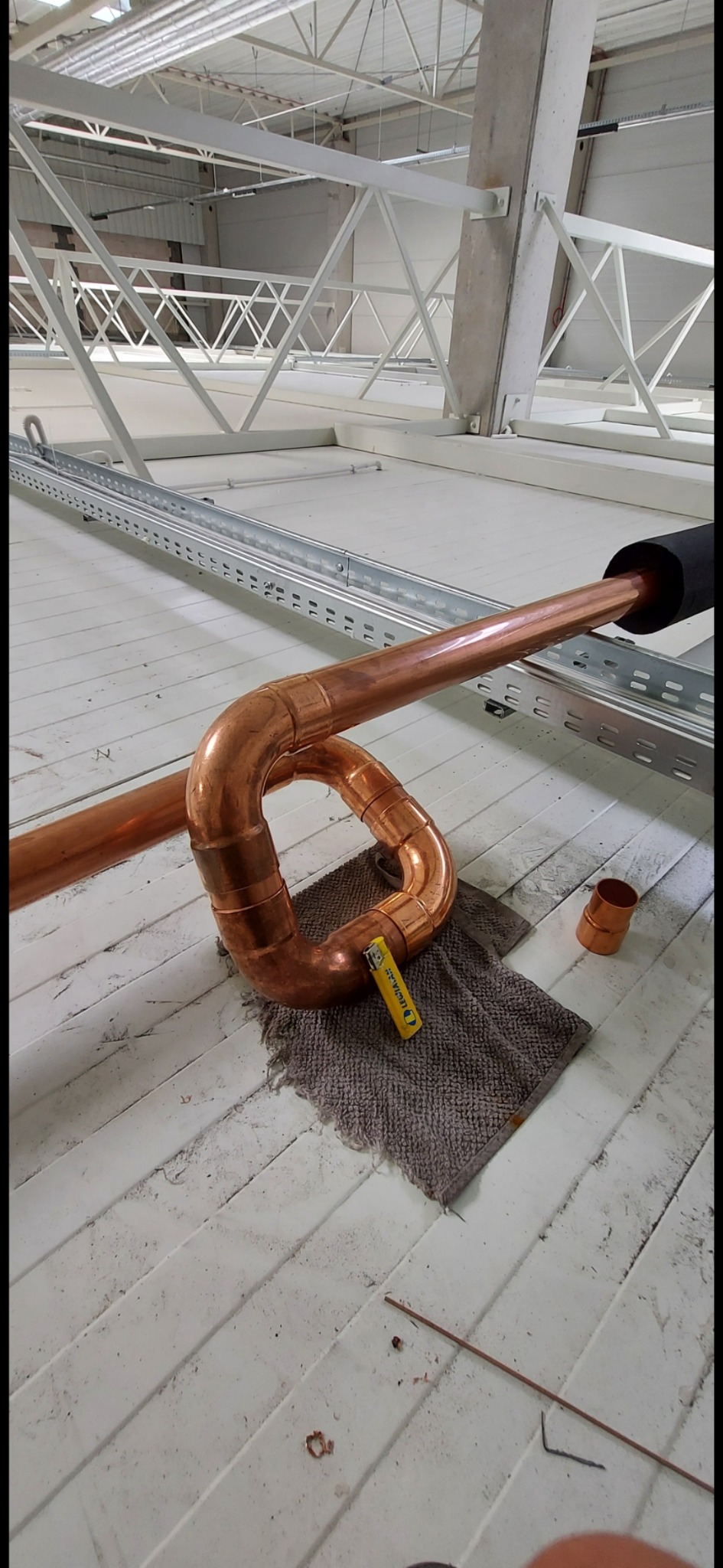 Miedziane rury w kształcie pętli, częściowo zaizolowane, leżące na szarej ścierce na tle sufitu hali z widoczną konstrukcją stalową