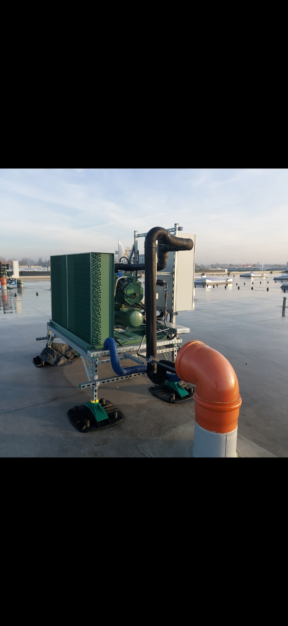 Agregat chłodniczy na dachu budynku, widoczne rury, przewody i elementy konstrukcyjne, tło stanowi niebo i odległe budynki.