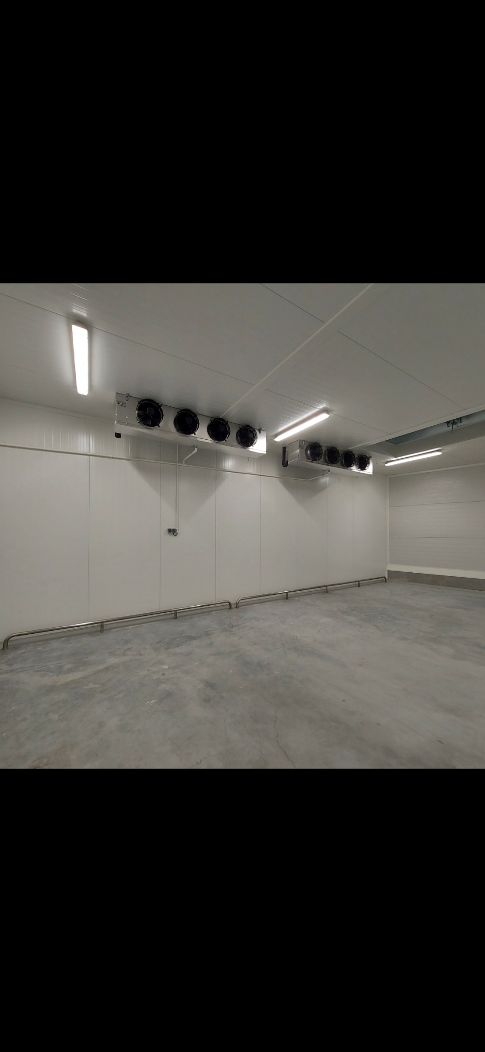 Wnętrze chłodni przemysłowej z zainstalowanymi agregatami chłodniczymi pod sufitem i zabezpieczeniami ścian. Widoczne białe panele ścienne i betonowa podłoga.