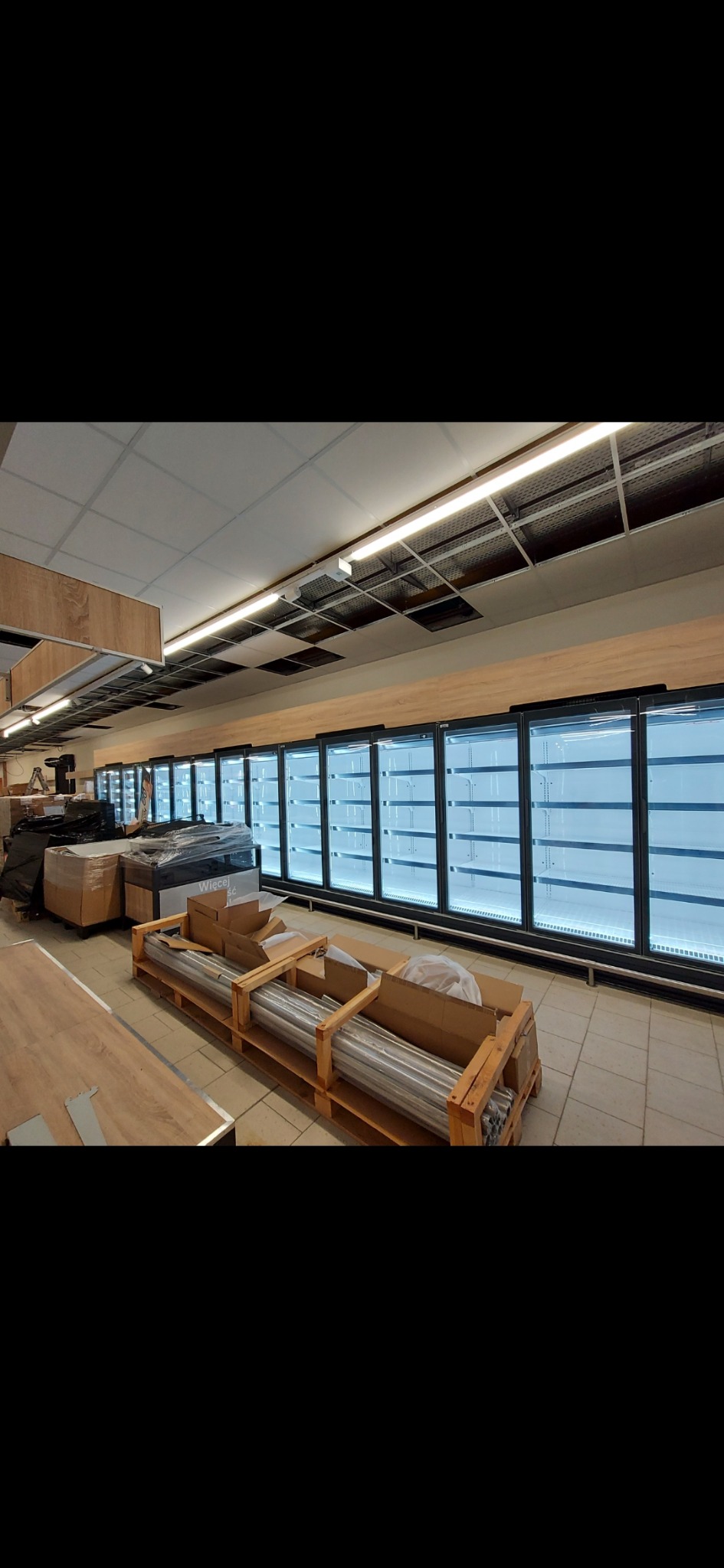 Montaż chłodniczych regałów sklepowych z przeszklonymi drzwiami, widoczne elementy instalacyjne w drewnianych paletach, surowy stan wykończenia wnętrza.